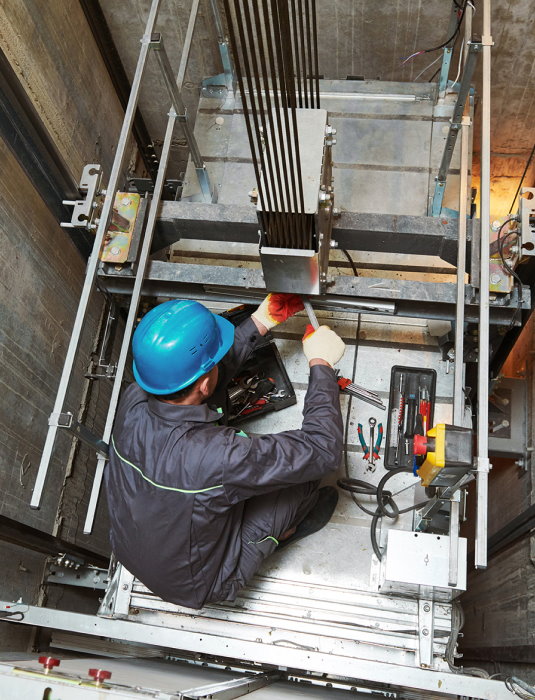 View from the top of an elevator shaft where a technician is working on the machinery below.