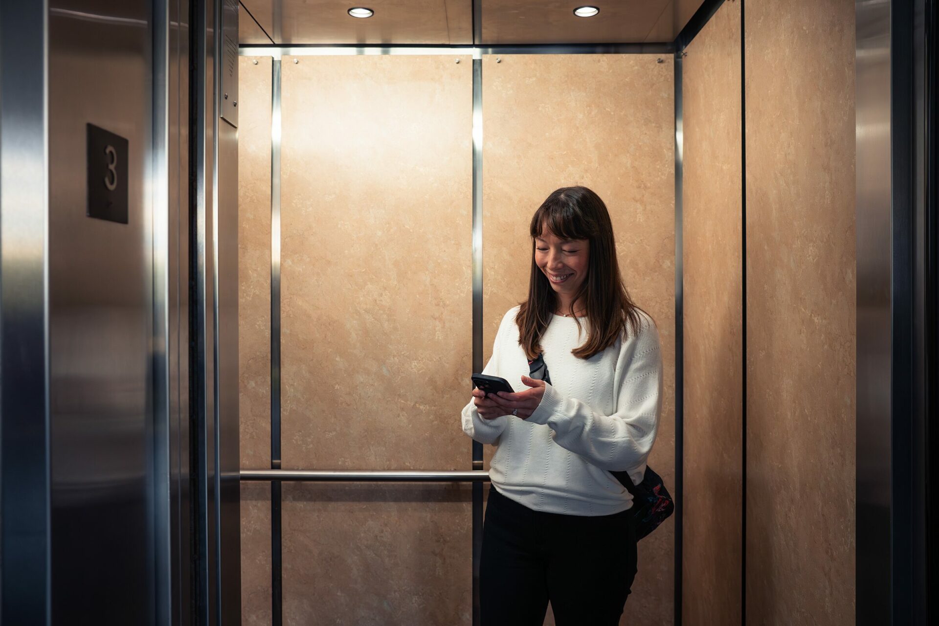 A smiling woman using a smartphone inside a modern elevator with a number three indicator above the door.