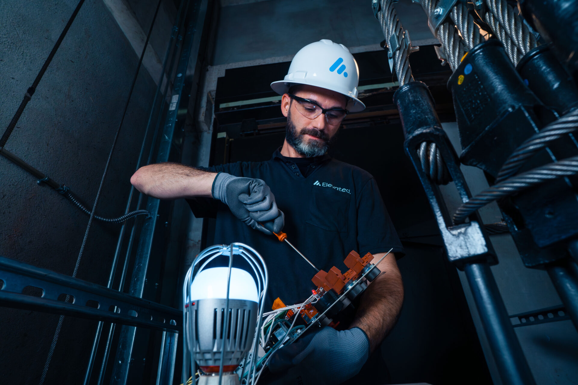 An elevator technician in a hard hat and glasses working on elevator wiring in a mechanical shaft.