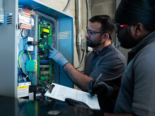 Two Elevated Facility Services technicians inspect an elevator control panel, reviewing its wiring and components during a service check.